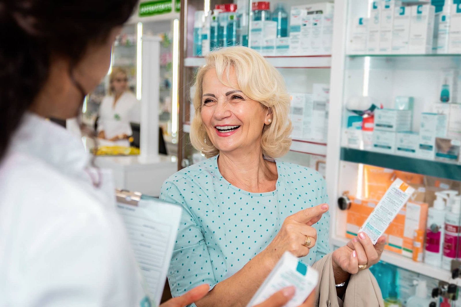 Una mujer rubia sonriente sostiene un producto de parafarmacia y señala su envase mientras consulta a una farmacéutica que sostiene una carpeta en una farmacia con estanterías blancas llenas de productos de cuidado personal.